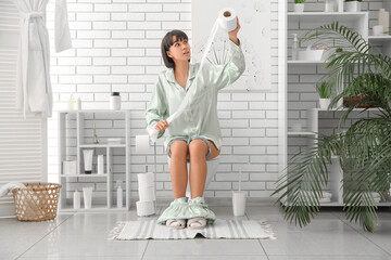 Young woman in pajamas with paper roll sitting on toilet bowl at home