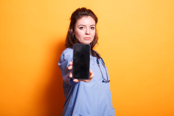 Caucasian woman in nurse uniform is holding smartphone with blank display. Medical assistant stands...