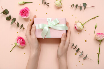 Female hands with gift box, beautiful roses and eucalyptus on pink background. International Women's Day