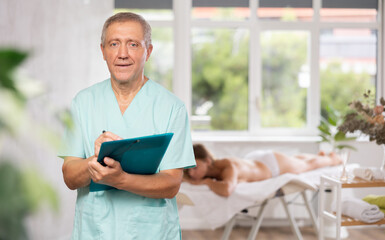 Positive old masseur posing with folder in hand in therapy room with patient lying on massage table