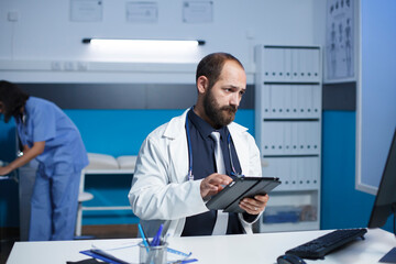Male doctor reading patient records while holding a tablet. In the clinic office, caucasian man wearing lab coat is using digital gadget and desktop pc to review medical information. - obrazy, fototapety, plakaty