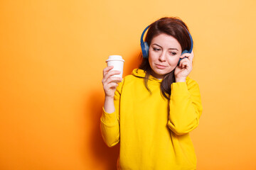 Portrait of female person with cup of coffee listening to music on headphones. Caucasian woman feeling relaxed and enjoying song on headset while holding hot beverage to drink. Young adult relaxing.