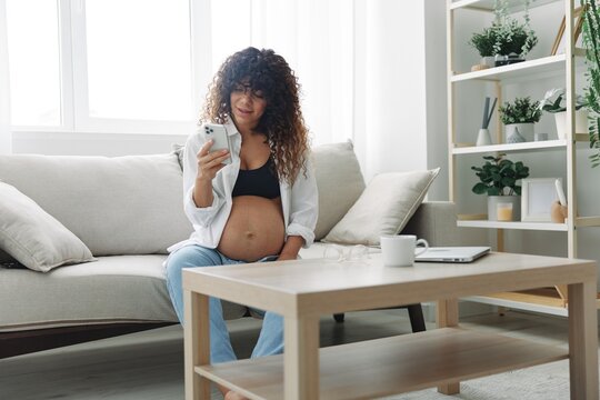 Pregnant woman blogger sits on the couch at home and takes pictures of herself on the phone, selfie and video call, online doctor consultation, pregnancy management, freelance work smiling