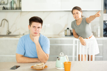 Upset young man sitting at the kitchen-table with his back to his wife disputing with him leaning on chair
