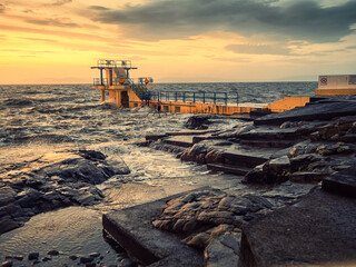Black rock diving board on Salthill beach in Galway city, Ireland. Warm sunset sky reflects in ocean water. Soft and dreamy cinematic look. Popular town landmark and tourist spot