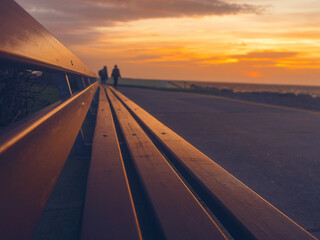 Sunrise sky reflects on long wooden bench, planks lead viewer to two person silhouette out of focus. Selective focus. Warm color and comfortable mood. . Salthill, Galway, Ireland - obrazy, fototapety, plakaty