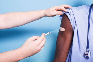Close-up of medical nurse being vaccinated in studio by doctor. Medic uses syringe with needle to administer vaccination injection to arm of african american healthcare specialist. - obrazy, fototapety, plakaty