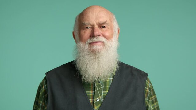 An agreeable elderly man in a checkered shirt and vest nods his head in a gesture of approval or affirmation, against a soothing turquoise background. Camera 8K RAW. 