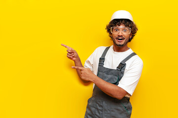 Young shocked Indian builder in hard hat and overalls points with hands to the side on yellow isolated background, surprised Indian foreman in uniform showing and advertising copy space in amazement - obrazy, fototapety, plakaty