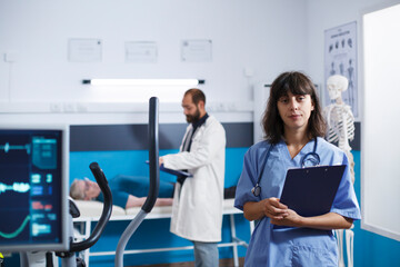 Medical assistant stands in physiotherapy office in this portrait. Looking at the camera, woman...