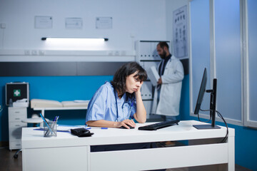 After hard day at hospital, a weary Caucasian nurse sitting at a desk with a desktop computer. Female practitioner caressing her face at a clinic office, looking exhausted.