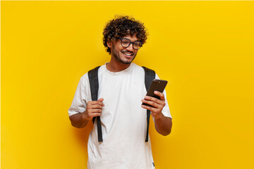 young indian guy student in a white t-shirt with glasses and with a backpack uses a smartphone and selects online on a yellow isolated background, a curly-haired man types a message on a mobile - obrazy, fototapety, plakaty