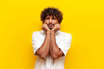 sad curly indian man thinking and looking up on yellow isolated background, young guy is bored and...