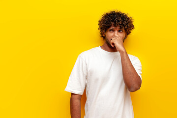 young curly indian man covering his nose with his hand and showing disgust on a yellow isolated...