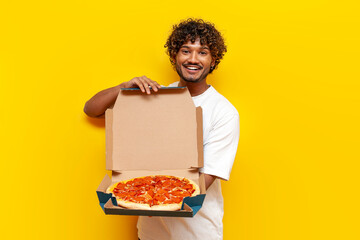 young indian man holding pizza box and showing on yellow isolated background, curly guy student showing and advertising fast food