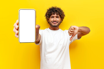 sad dissatisfied Indian man showing blank smartphone screen and dislike on yellow isolated background, curly guy advertising phone online and showing bad review - obrazy, fototapety, plakaty