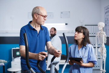 Physiotherapy being administered to patient as he pedals on stationary bike, with assistance of female nurse dressed in scrubs. Healthcare assistant holds tablet to relay medical results to old man.