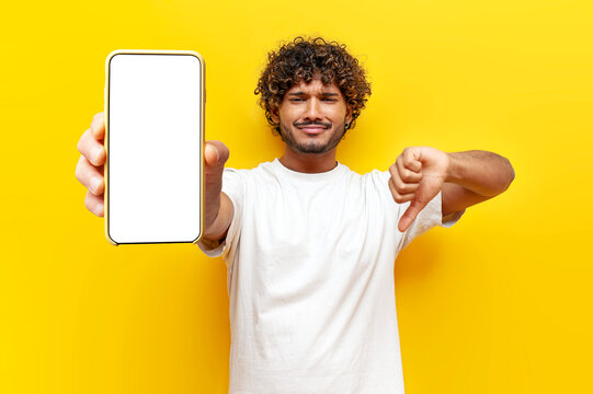 sad dissatisfied Indian man showing blank smartphone screen and dislike on yellow isolated background, curly guy advertising phone online and showing bad review