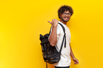 young indian guy student in a white t-shirt with glasses and with backpack points back on yellow isolated background, curly man goes to college to study and points at the copy space from behind - obrazy, fototapety, plakaty