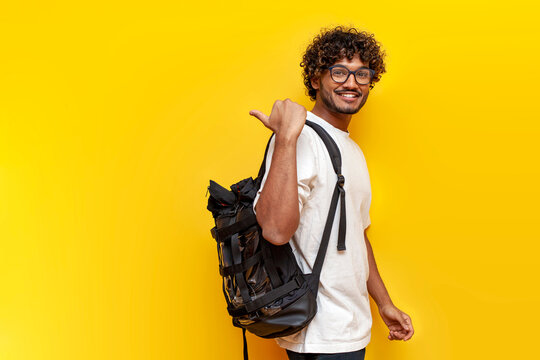 Young Indian Guy Student In A White T-shirt With Glasses And With Backpack Points Back On Yellow Isolated Background, Curly Man Goes To College To Study And Points At The Copy Space From Behind