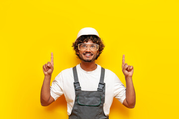 young indian male builder in hard hat and overalls points with his hands up on a yellow isolated...