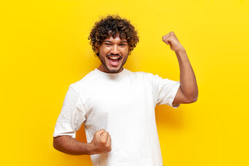 cheerful indian man celebrating victory and good luck with raised hands on yellow isolated background, curly guy wins and rejoices at success - obrazy, fototapety, plakaty