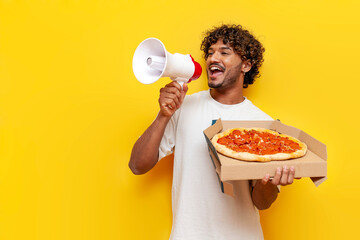 young Indian man holding a delicious pizza and announcing information into a loudspeaker on a yellow isolated background, a curly-haired guy shouts into a megaphone and advertises fast food - obrazy, fototapety, plakaty