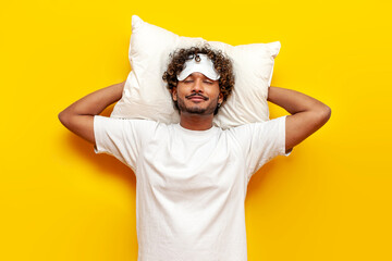 young cheerful indian man in pajamas and sleep mask lies on a soft pillow sleeping and resting on a yellow isolated background, curly guy getting ready for bed