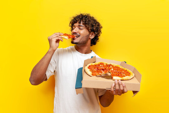 young indian man eating delicious pizza on yellow isolated background, curly guy student holding pizza box and biting a piece of fast food