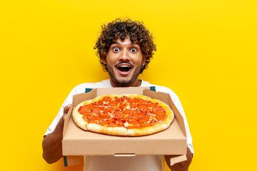 young shocked indian man holding a box of delicious pizza and wondering on yellow isolated...