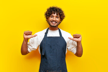 cheerful indian male waiter in an apron points to himself on a yellow isolated background, indian...