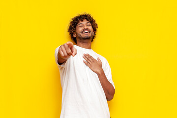 young indian man in a white t-shirt mocks and jokes and points his finger forward on a yellow isolated background, curly-haired guy laughs and ridicules and mocks - obrazy, fototapety, plakaty