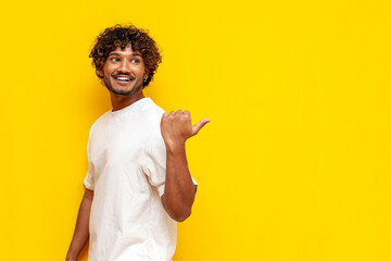 young cheerful indian man in white t-shirt pointing with hand back and smiling on yellow isolated...