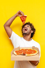 young indian man holding a pizza box and eating a piece on a yellow isolated background, curly guy...