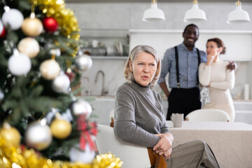 Elderly woman sad while sitting at table in kitchen with happy couple of man and woman