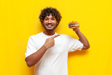 young indian man holding tasty burger and pointing with hand on yellow isolated background, curly...