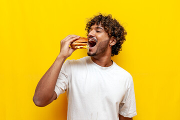 young indian man eating a big tasty burger with his mouth wide open on a yellow isolated background, curly guy student eating and advertising fast food - obrazy, fototapety, plakaty