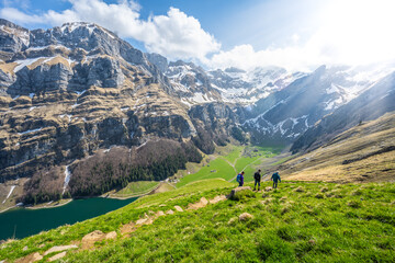 Tourists walk along a hiking trail through a green meadow with an alpine lake and a steep, rocky mountain formation in the background. Seealpsee, Appenzell, Switzerland, Europe.