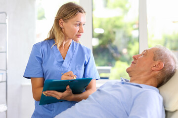 Female doctor holding document folder interacting attentively with elderly man lying on couch in...