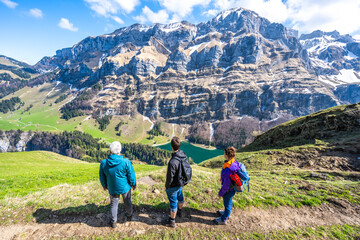 Fototapeta na wymiar Tourists on hiking trail at a green meadow overloop the alpine lake and rocky mountain formation in the background. Seealpsee, Appenzell, Switzerland, Europe.