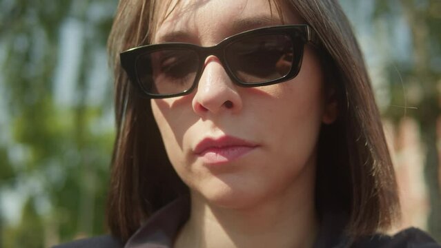 Portrait of young brunette woman in black sunglasses in the park. Wind in brown hair close-up, sunny day outdoors