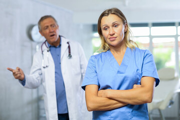 Frustrated female doctor standing with crossed arms in medical office on background with disgruntled senior colleague reprimanding her