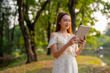Naklejka premium Beautiful Young Asian Woman Finds Serenity, Nature, and Inspiration While Holding a Digital Tablet in the Sunset Breeze at the Park