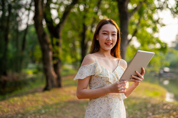 Naklejka premium Beautiful Young Asian Woman Finds Serenity, Nature, and Inspiration While Holding a Digital Tablet in the Sunset Breeze at the Park