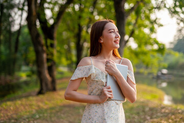 Beautiful Young Asian Woman Finds Serenity, Nature, and Inspiration While Holding a Digital Tablet in the Sunset Breeze at the Park