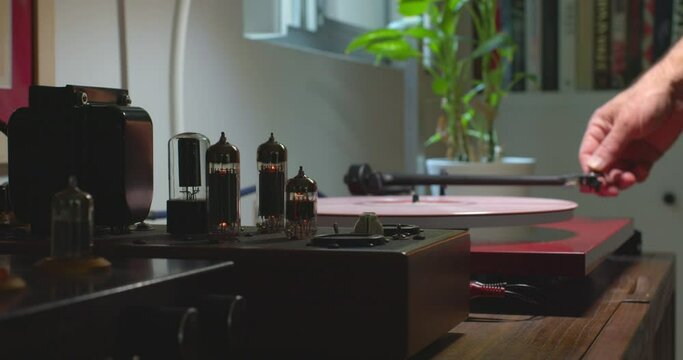 Camera on slider smoothly pushing past tube amplifier to turntable with man placing a stylus on pink colored vinyl record.