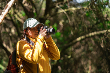 Woman walking with a red backpack and blue cap taking photos in the middle of a forest during a sunny day