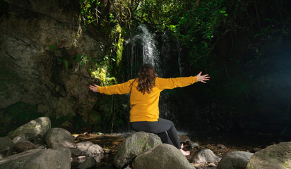 Woman dressed in a yellow jacket and black pants sitting next to a waterfall, with her arms open, in the middle of a forest during a sunny day