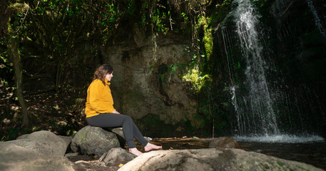 Woman dressed in a yellow jacket and black pants sitting next to a waterfall in the middle of a forest during a sunny day