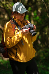 Woman walking with a red backpack and blue cap reviewing the photos on her camera in the middle of a forest during a sunny day